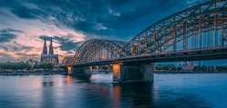 Panoramic view of illuminated bridge over Rhine river whith trains and tourists passing by and the Cologne Cathedral in the evening at the blue hour.