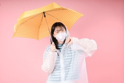Young asian woman isolated on pink background showing thumb down wears protective mask to reduce breathing pollutants, wears raincoat, hides under umbrella. Air pollution concept