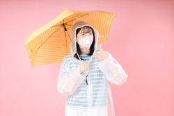 Asian girl raising thumbs up, wears protective mask to reduce breathing pollutants, wears raincoat, hides under umbrella. Air pollution concept