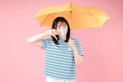 Young asian woman isolated on pink background showing thumb down wears protective mask to reduce breathing pollutants, hides under umbrella. Air pollution concept