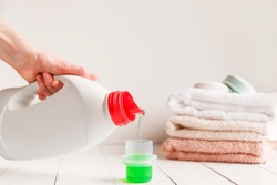Close up of female hands pouring liquid laundry detergent into cap on white rustic table with towels on background in bathroom.