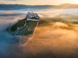 Aerial view of Spiš Castle, surrounded by fog, lit by morning sun. Extensive castle ruins above the valley against mountains in background. Magic colors, orange colored mist and blue shadows. Slovakia
