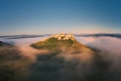 Aerial view of Spiš Castle surrounded by fog, lit by orange sun. Castle ruins, standing on a   white rock above the valley against Tatra mountains in background.  UNESCO site. Traveling concept. 