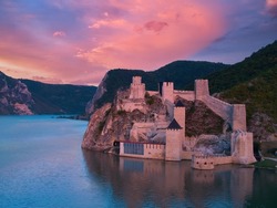 Aerial, across lake view of the medieval fortress Golubac over  Danube river. Fortress towers illuminated by pink light. Sunset, pink and red clouds sky. Outdoor and traveling theme. Serbia.