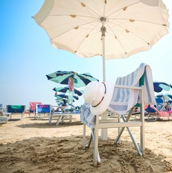 Chair with white straw hat and striped towel under a beach umbrella on a sunny summer day. In the background, the beach equipped with sunbeds and umbrellas along the Adriatic Sea in Italy.