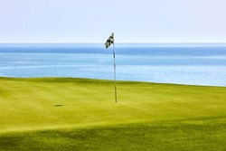 Pole with black and white checkered flag in the center green on a golf course on a summer day. In the background the sky and the blue sea.