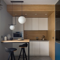Small and stylish kitchen with modern white furniture, wooden walls and table with two, black chairs under modern lamp