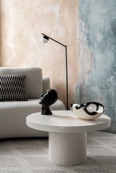 Modern, white and round coffee table with decorative black sculpture and bowl in stylish room with beige sofa, black lamp and colorful concrete wall