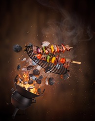 Kettle grill with hot briquettes, cast iron grate and tasty skewers flying in the air. Freeze motion barbecue concept.