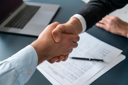 Close up of two businessmen shaking hands in blurred office with computer and contract on table. Concept of partnership and communication