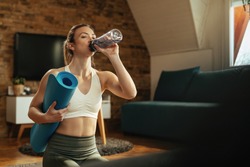 Thirsty athletic woman drinking water from a bottle after sports training at home. 