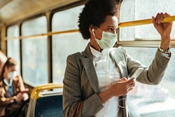 Black businesswoman with protective face mask using smart phone and looking through the window while commuting by bus.  