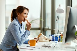Young businesswoman using cell phone while eating a cookie at her office desk. 