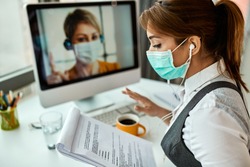 Businesswoman with face mask going through reports while having online meeting with her colleague in the office. 