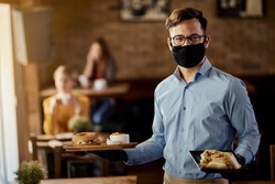 Young happy waiter wearing protective face mask while serving food in a restaurant. 