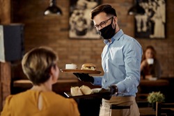 Happy waiter wearing protective face mask and gloves while bringing food to a customer in a pub. 