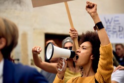 Young African American woman with raised fist shouting through megaphone while being on anti-racism protest. 