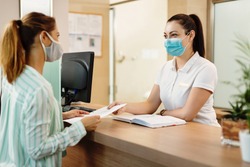 Female receptionist taking application form from her customer at health spa check in counter. They are wearing protective face masks due to COVID-19 pandemic. 