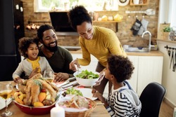 Happy African American woman having Thanksgiving lunch with her family and serving salad at dining table. 