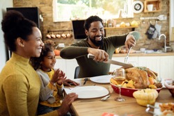 Happy African American man carving roasted turkey while having Thanksgiving lunch with his family at dining table. 