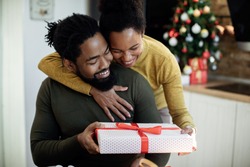 Happy black woman embracing her husband and giving his a gift on Christmas day at home. 