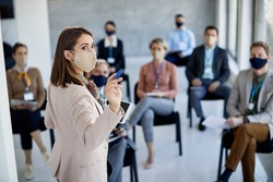 Female executive manager with face mask talking to group of business people during seminar in board room.