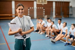Young happy female coach looking at camera while having PE class with elementary students at school gym. 