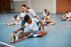 Black school boy stretching on the floor with help of PE teacher while warming up exercise class. 