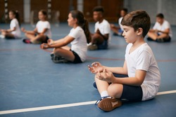 Group of elementary students doing breathing exercise while practicing yoga during physical education class at school. Focus is on a boy in foreground. 