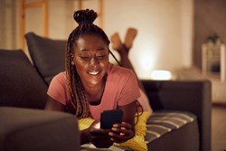 Happy black woman using smart phone and text messaging while relaxing at night in the living room. 