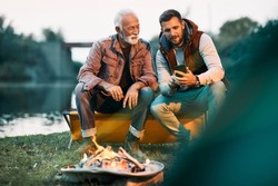 Happy senior man and his son using mobile phone while camping and relaxing by campfire. Copy space.