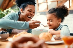 Happy black mother having fun and feeding her daughter while having breakfast at dining table.