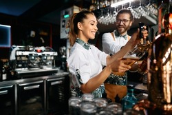 Young happy bartenders pouring beer from a beer tap while working in a pub. Focus is on woman.
