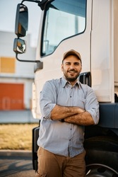 Happy truck driver standing with arms crossed in front of his truck and looking at camera.
