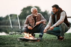 Happy mature man and his adult son talking while relaxing by campfire during their fishing day.