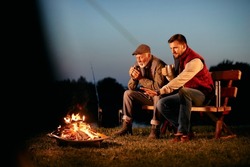 Senior mand and his adult son having a drink while relaxing by bonfire at dusk during their camping weekend.  Copy space.
