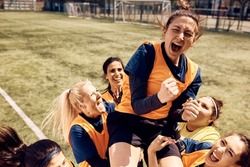 Cheerful team of female soccer players celebrating victory and carrying on of teammates who is shouting out of joy on stadium.