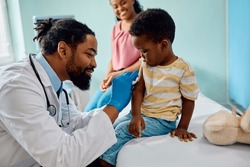Caring African American pediatrician preparing arm of a small boy for vaccination at doctor's office.