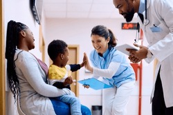 Small African American kid giving high five to pediatric nurse while being with his mother at doctor's office.