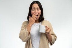 Asian woman wearing domestic clothes holding cup of coffee in hand and yawning at morning. Daily morning routine and start of a new day concept 
