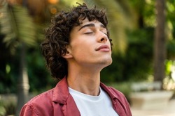 Tranquility. Portrait of a happy caucasian curly man relaxing with closed eyes while standing at the street at the park. Stock photo 