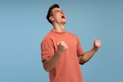 Portrait of happy screaming handsome young man standing with suprised face and rejoicing his victory. Indoor studio shot, isolated on blue background 