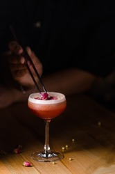 Classic cocktail glass on a wooden table in a nightclub restaurant. Alcoholic cocktail of pink color with a rose, close-up. Modern alcoholic drink. Bartender's hands decorate a cocktail