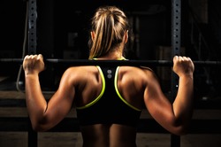Back view young adult girl doing barbell squats in gym. Woman with muscular body doing lifting exercise.