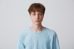 Close up face of a young man without emotions. Beautiful emotionless guy in a blue t-shirt looking to the camera, isolated over white background