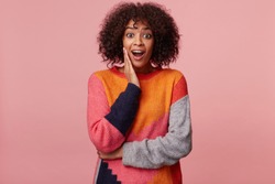 Surprised African American woman with an afro hairstyle with amazement looks camera, palm holds her cheek, feels impressed, looks affected excited overwrought, isolated on a pink background