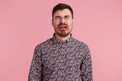 Portrait of a sad crying bearded man in glasses, wearing in colorful shirt, looks unhappy and upset. Isolated over pink background, people and emotions concept.