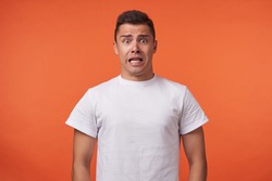 Frightened young brown-eyed brunette man with short haircut rounding amazedly his eyes while looking scaredly at camera, standing over orange background