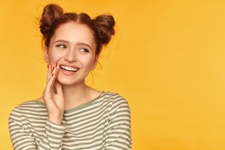 Young lady, pretty ginger woman with two buns and healthy skin. Smile and touch corner of a mouth. Wearing striped sweater and watching to the right at copy space, closeup over yellow background