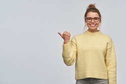 Smiling, beautiful girl with blond hair gathered in bun. Wearing yellow sweater and eyewear. Pointing with thumb to the left at copy space. Watching at the camera, isolated over white background